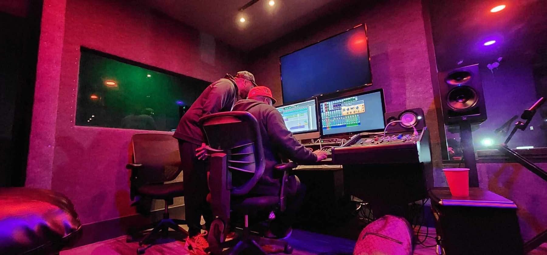 Engineered music session in the control room — DAW open, booth lit beyond the glass